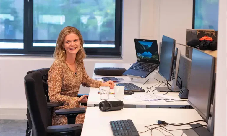 Flexible office workspace with a woman smiling at her desk, multiple computer screens, and a laptop, showcasing a modern work environment and career opportunities at Keylane.